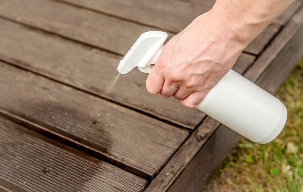 Person Spraying a White Bottle onto A Weathered Wooden Deck Outdoors — My Habitat Products in Malanda, QLD