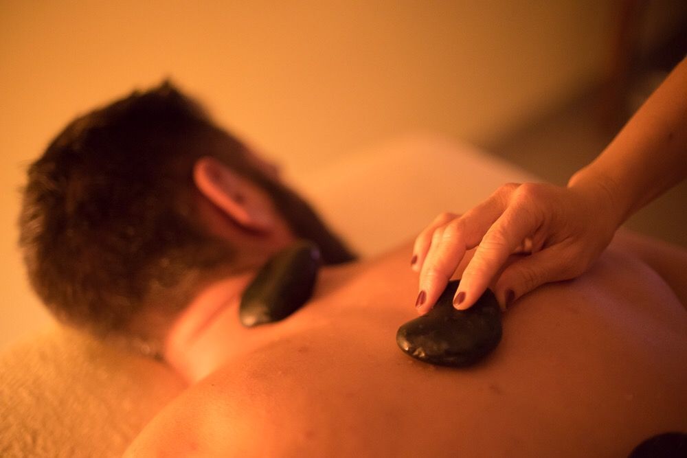 Uomo che riceve un massaggio con pietre calde. Una mano gli appoggia una pietra liscia e scura sulla schiena. Luce soffusa e calda.
