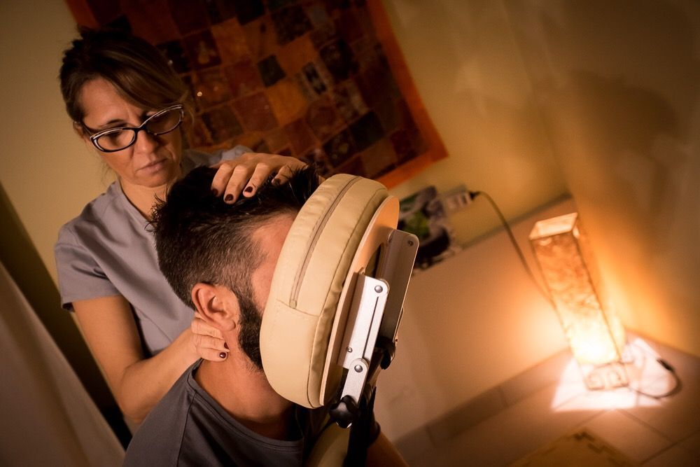 Donna seduta che esegue un massaggio a una persona; in una stanza scarsamente illuminata, con una lampada e opere d'arte.