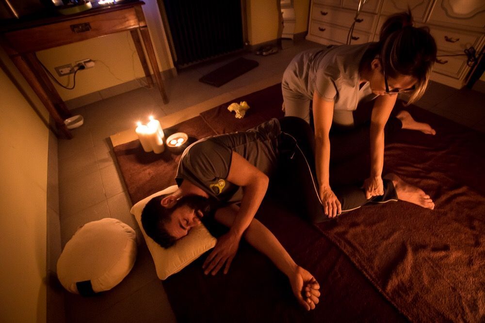 Uomo che riceve un massaggio su un tappetino. Il terapista è inginocchiato e lavora sulla gamba. Le candele bruciano debolmente.