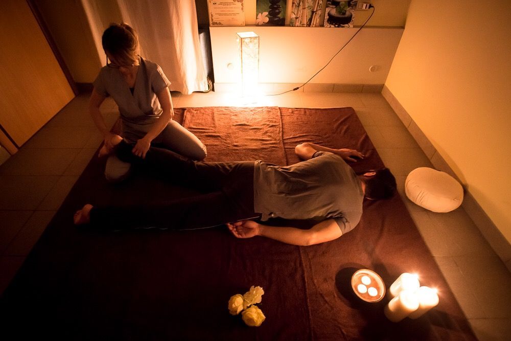Persona che riceve un massaggio sul tappetino, terapista inginocchiato. Candele, luci soffuse, interni.