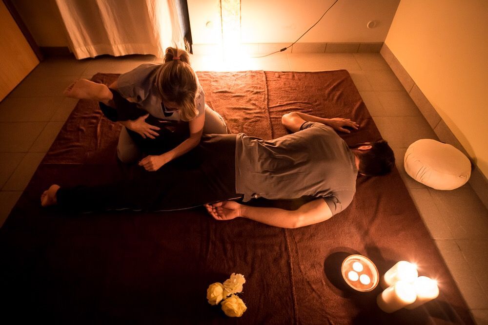 Donna che esegue un massaggio thailandese su un uomo sdraiato su un materassino. Candele e asciugamani in una stanza scarsamente illuminata.