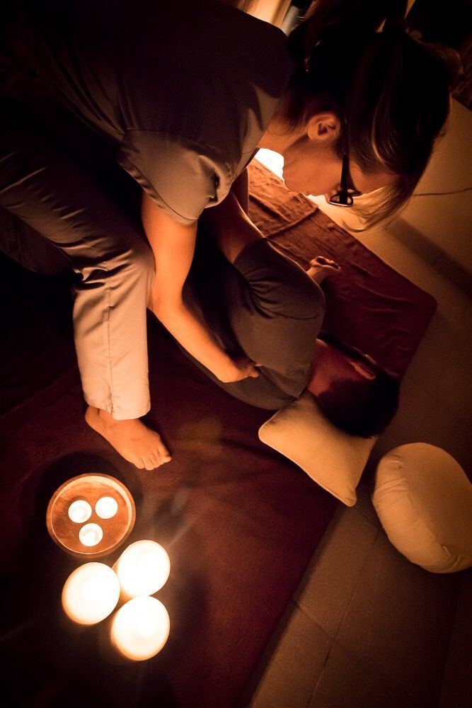 Persona che esegue un massaggio in una stanza scarsamente illuminata, con candele sul pavimento e una persona sdraiata.