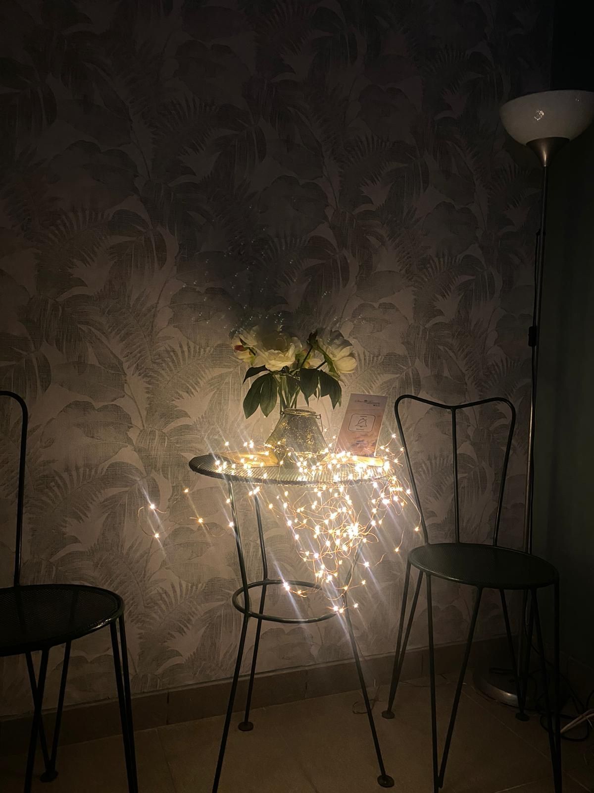 Un tavolino con lucine, fiori e due sedie nere, illuminato da una lampada da terra contro una parete decorata.