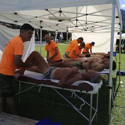 Uomini che ricevono massaggi durante un evento all'aperto sotto una tenda bianca. I massaggiatori indossano magliette arancioni.
