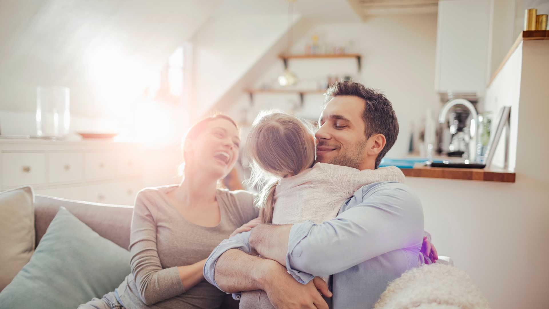 Family of three hugging on a couch, smiling in a sunlit living room.