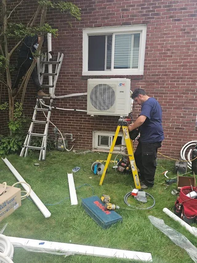 A technician installs an outdoor HVAC unit on a brick wall, standing on a yellow ladder with tools scattered on the grass.