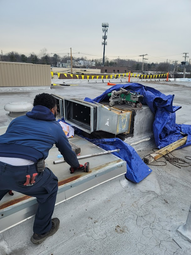 A worker in a blue hoodie kneels on a flat rooftop, installing metal ductwork covered by a blue tarp.