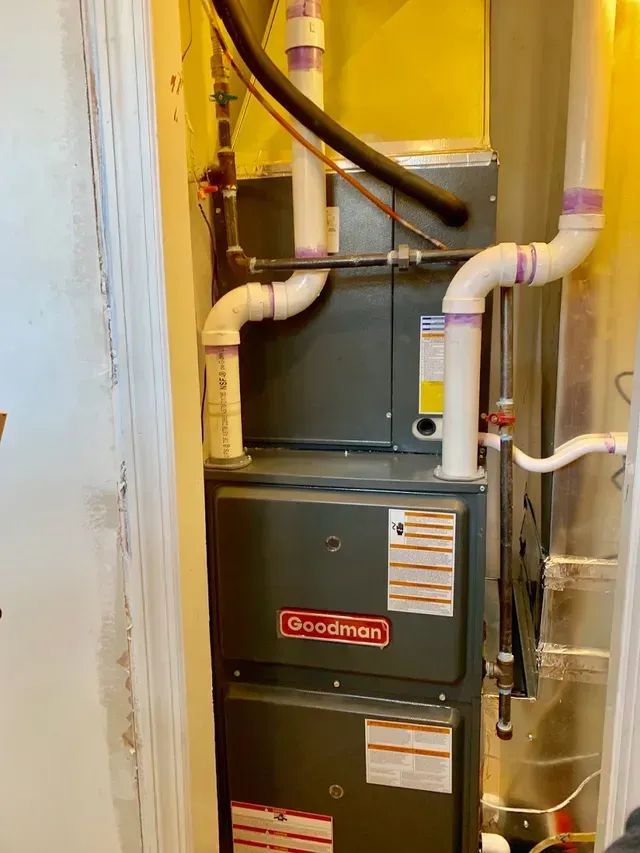 A Goodman furnace and air handler unit with white PVC intake and exhaust pipes, installed in a narrow closet.