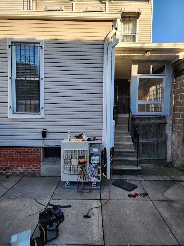 A technician's HVAC unit stands on a patio with tools and gauges attached, next to a house with beige siding and steps.