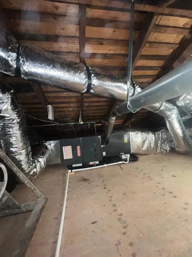 HVAC air handler unit installed in an attic with insulated ductwork and a white PVC condensate drain pipe on the floor.