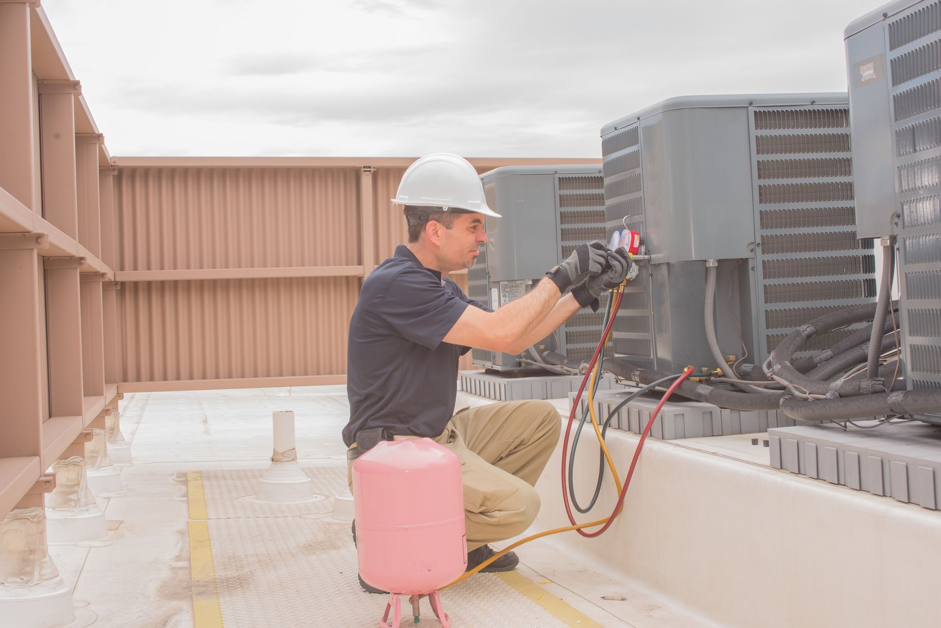 A technician in a hard hat kneeling on a rooftop, using gauges to service an outdoor HVAC unit.