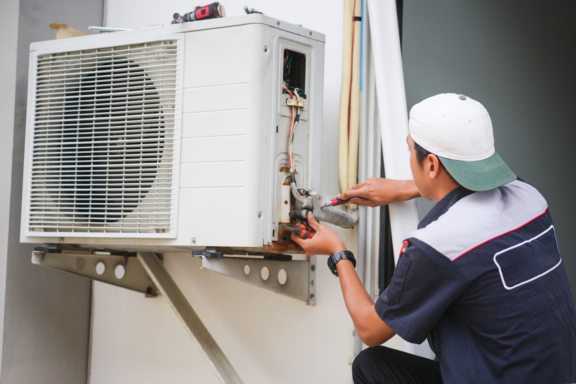 A technician in a uniform and cap repairs an outdoor air conditioning unit mounted on a wall.