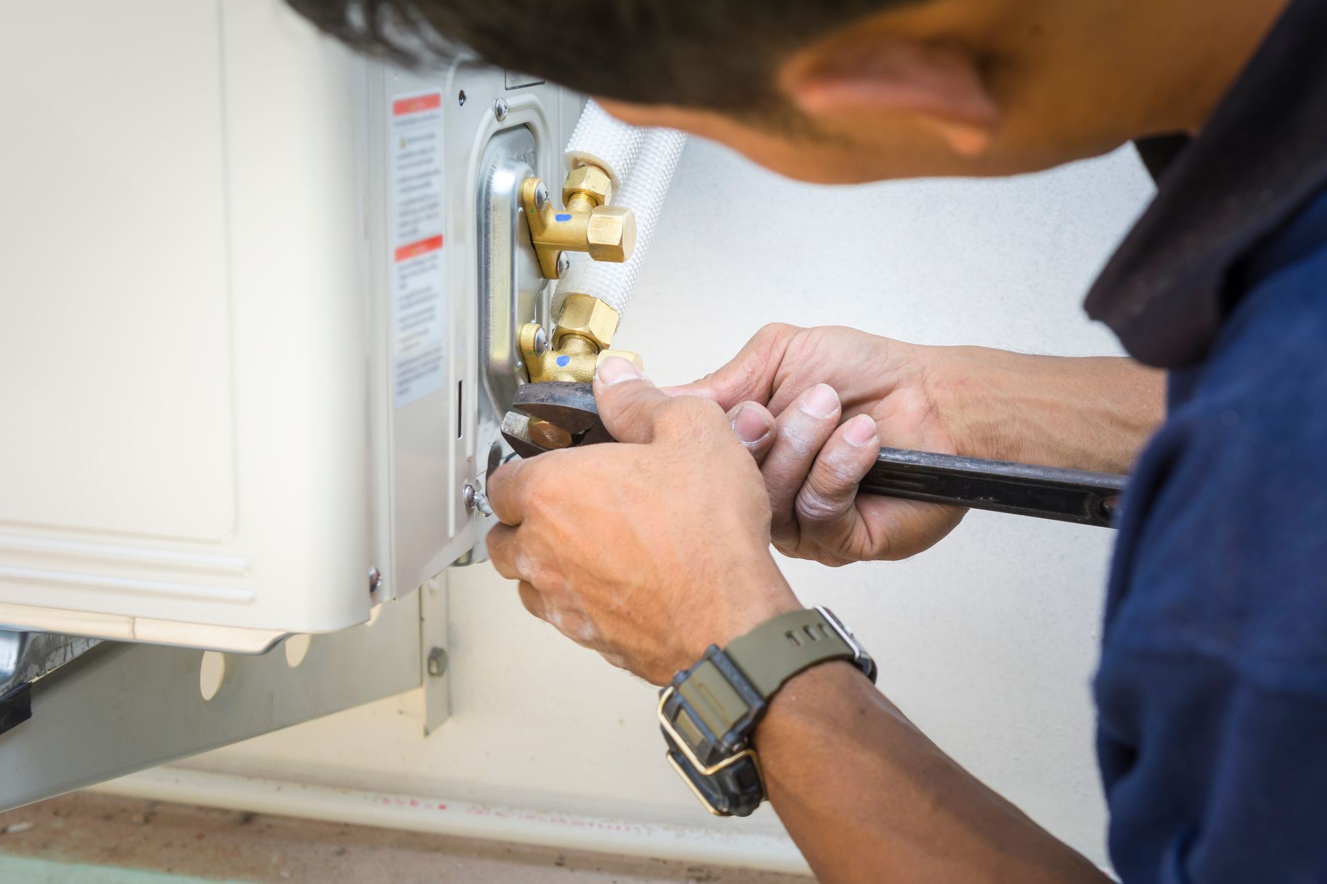 A person uses a wrench to tighten a brass valve fitting on the side of an outdoor air conditioning unit.