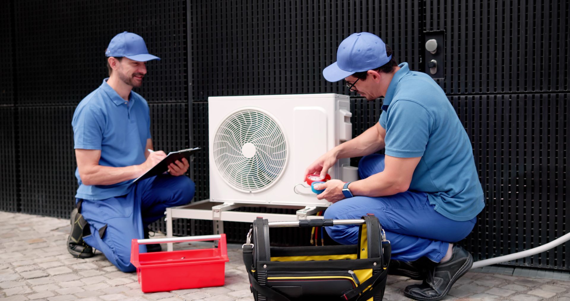 Two workers in blue uniforms kneeling outdoors to repair and inspect an air conditioning unit with tools and a clipboard.