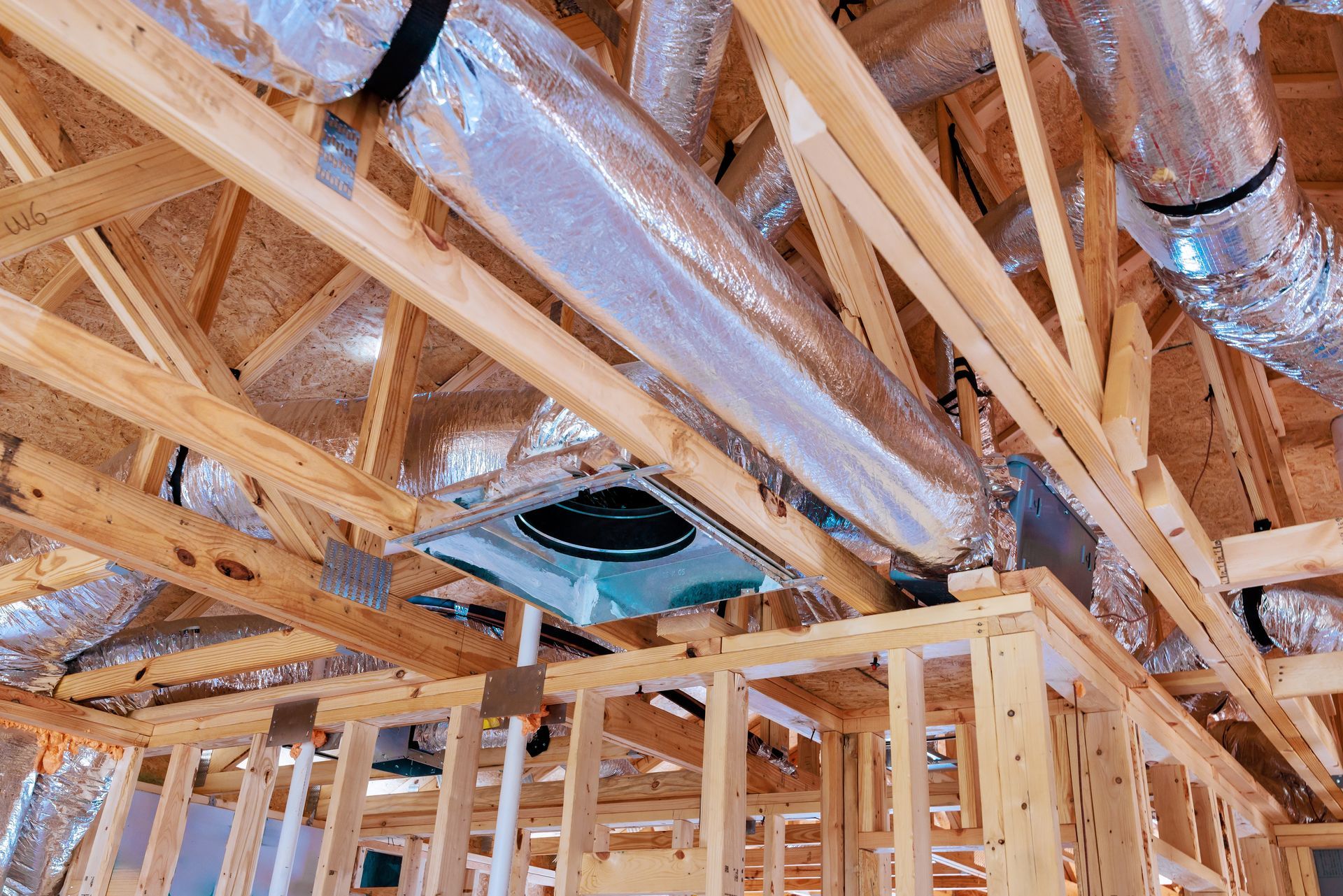 Unfinished wooden ceiling trusses with silver insulated HVAC ducts running through them in a building under construction.