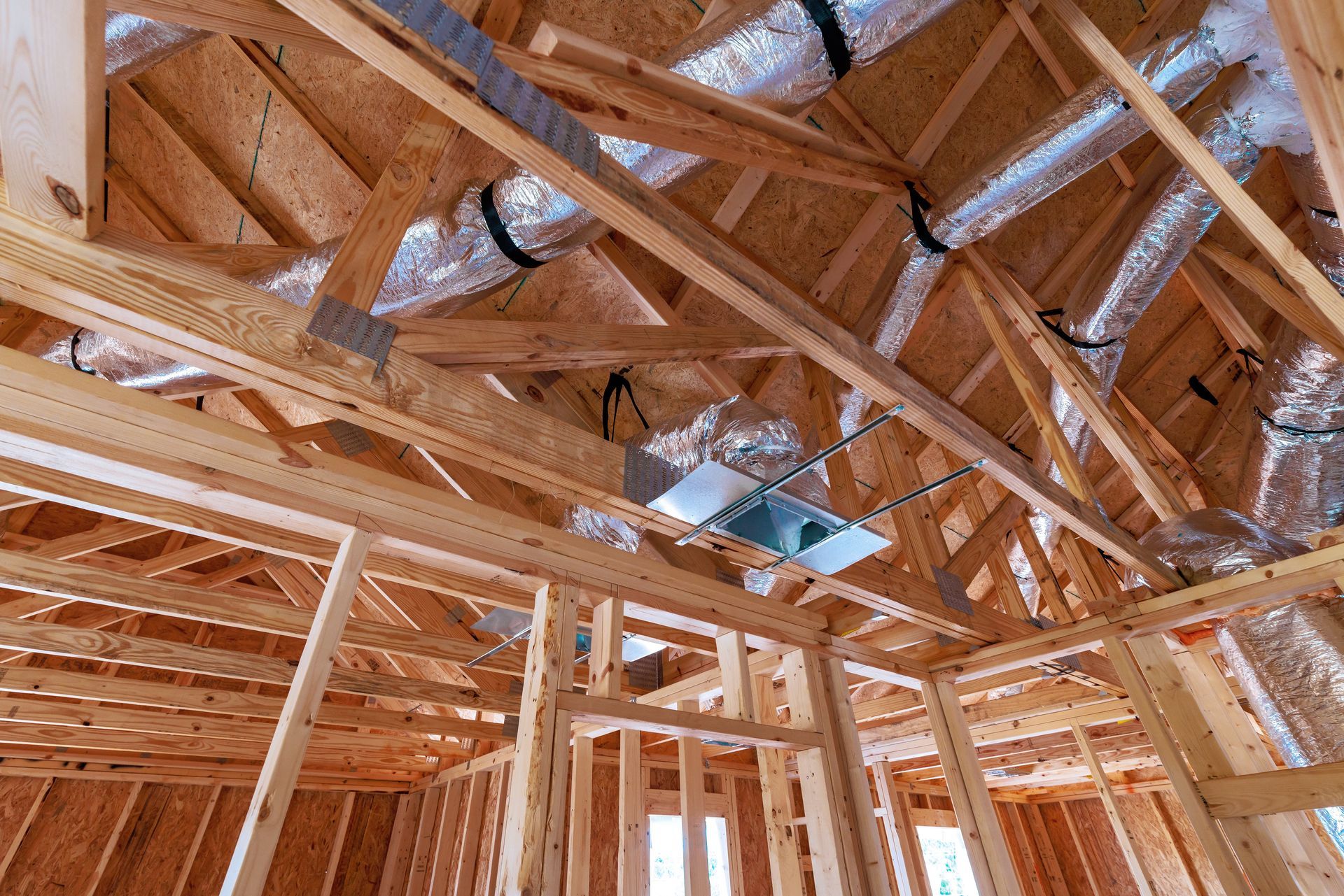 The interior framework of a house under construction, showing wooden roof trusses, support beams, and metallic ductwork.