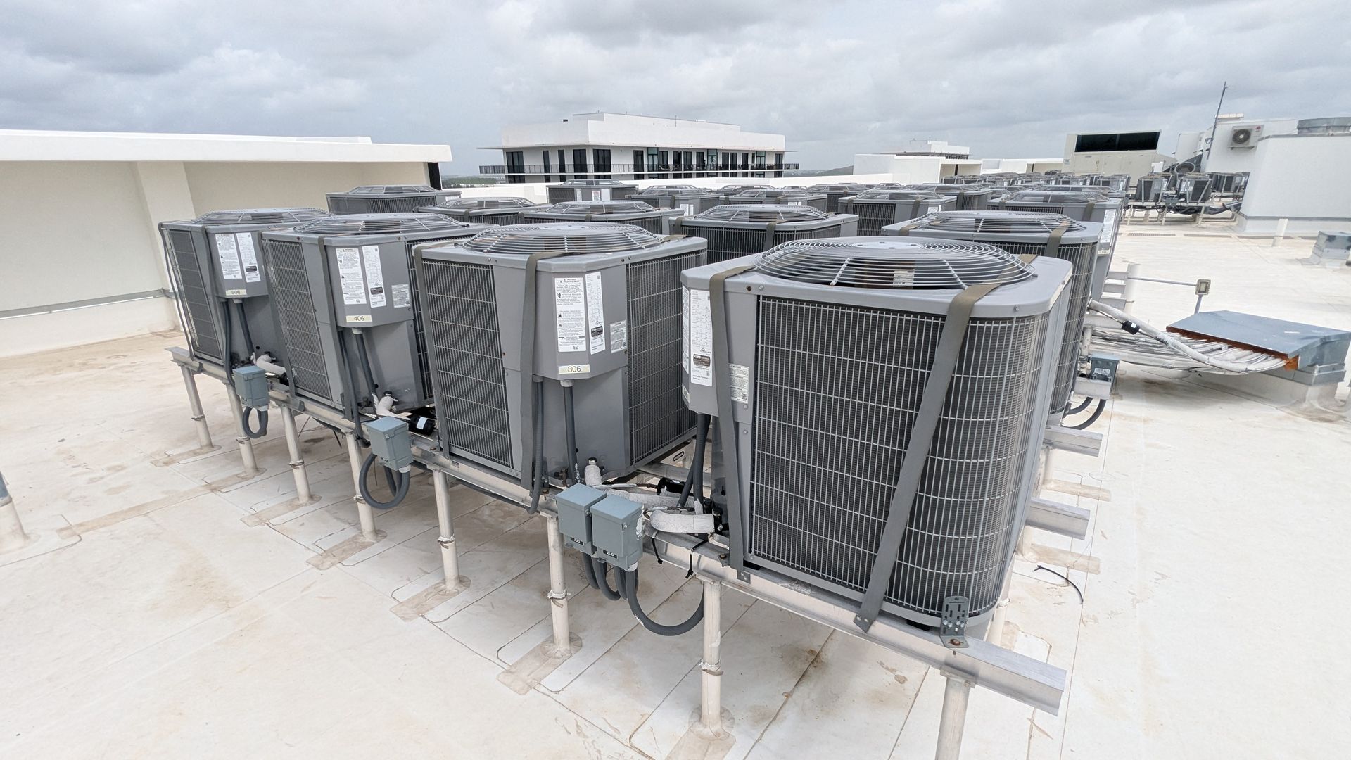 Multiple gray HVAC air conditioning units arranged in rows on a flat commercial rooftop under a cloudy sky.