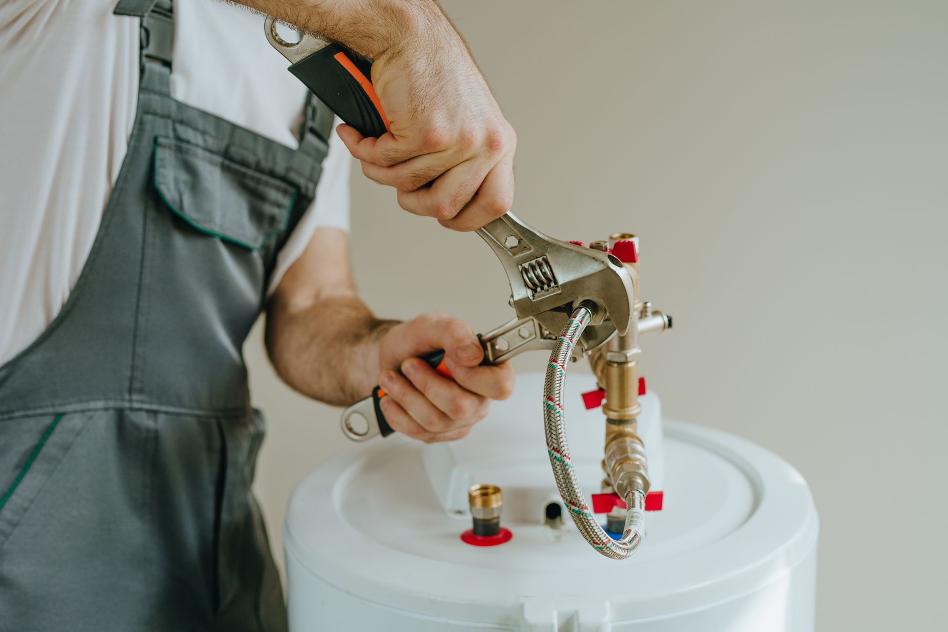 A worker in grey overalls uses an adjustable wrench to tighten a metal hose connection on a white water heater.