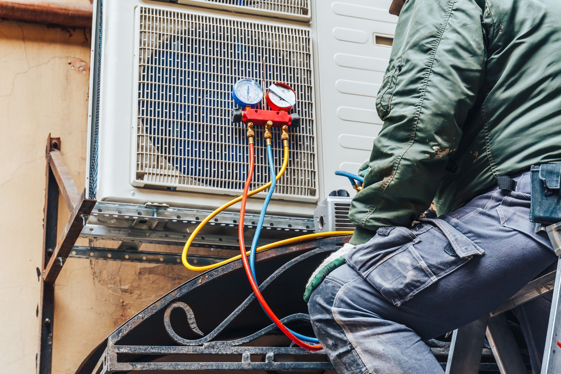 A technician in work clothes services an outdoor air conditioning unit using a manifold gauge set with colored hoses.