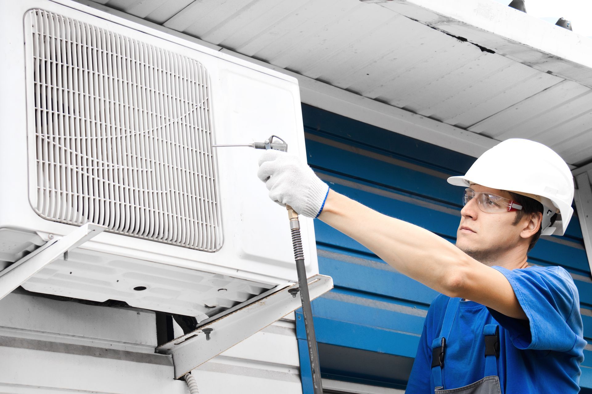 A technician in a hard hat and protective glasses uses an air nozzle to clean an outdoor air conditioning unit.