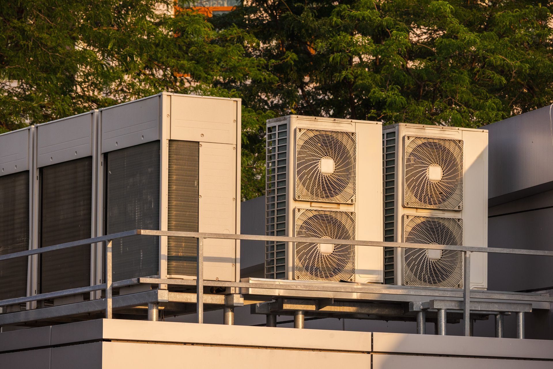 Industrial HVAC cooling units on a rooftop with a metal railing and green trees in the background under warm sunlight.