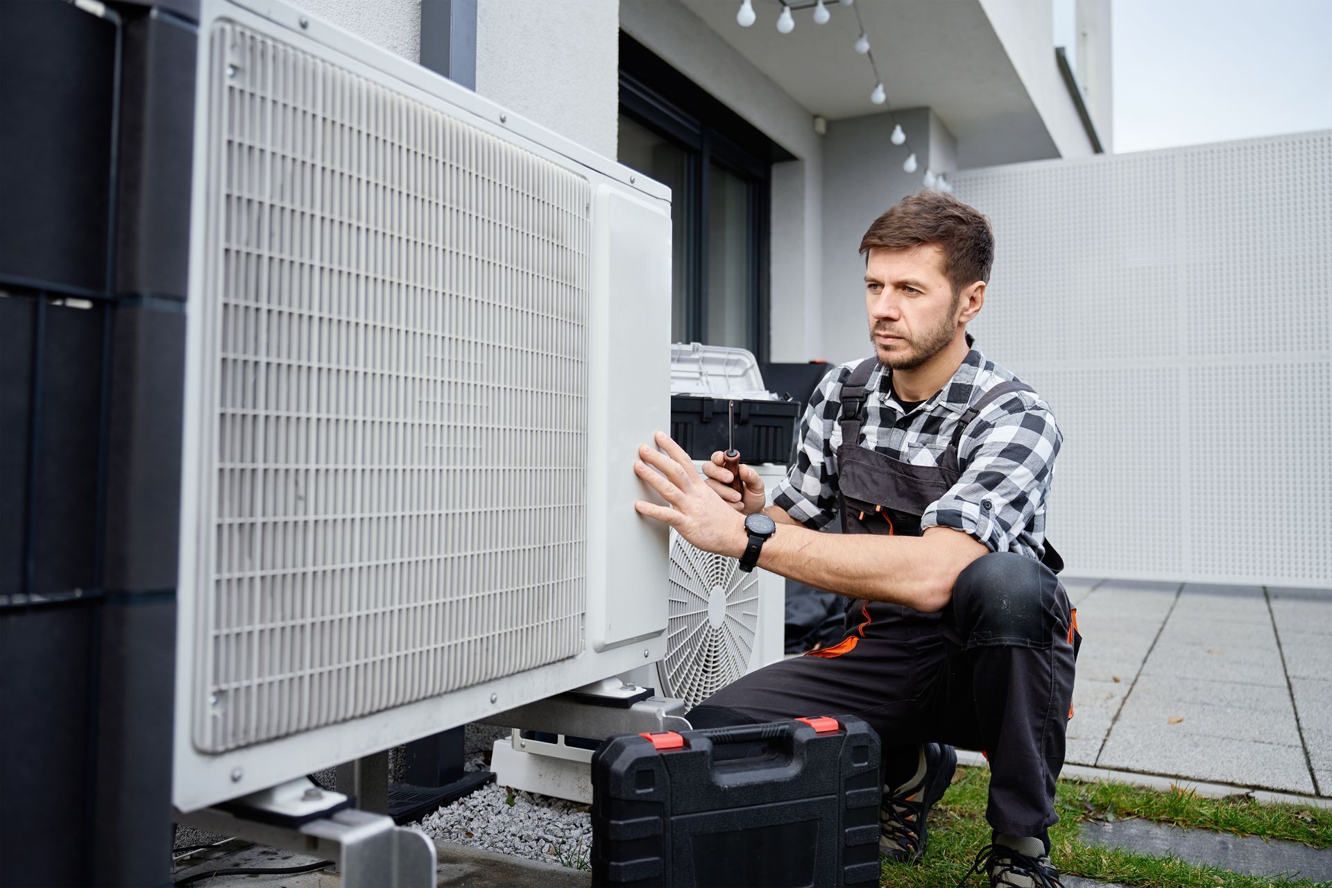 A technician in work coveralls kneels outdoors, inspecting an air conditioning unit with a tool in hand.