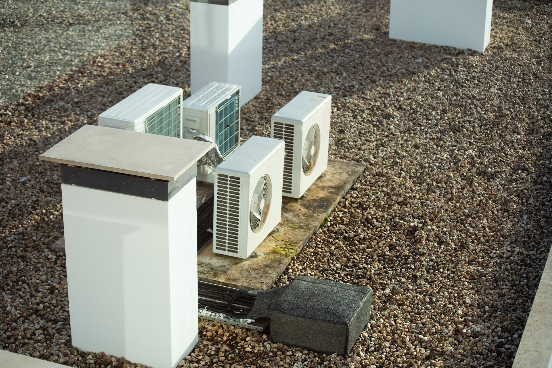A cluster of outdoor air conditioning condenser units on a gravel surface next to white, block-shaped pedestals.