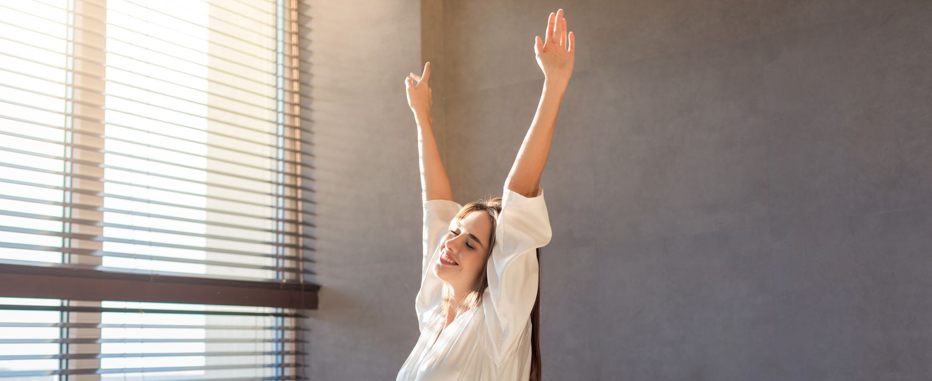 A person in a white shirt stretches with arms raised, smiling near a window with horizontal blinds in a sunlit room.
