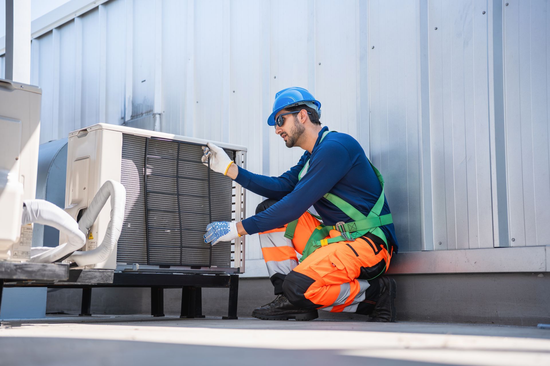 A technician in safety gear kneels on a rooftop, inspecting the condenser coils of an outdoor HVAC unit.