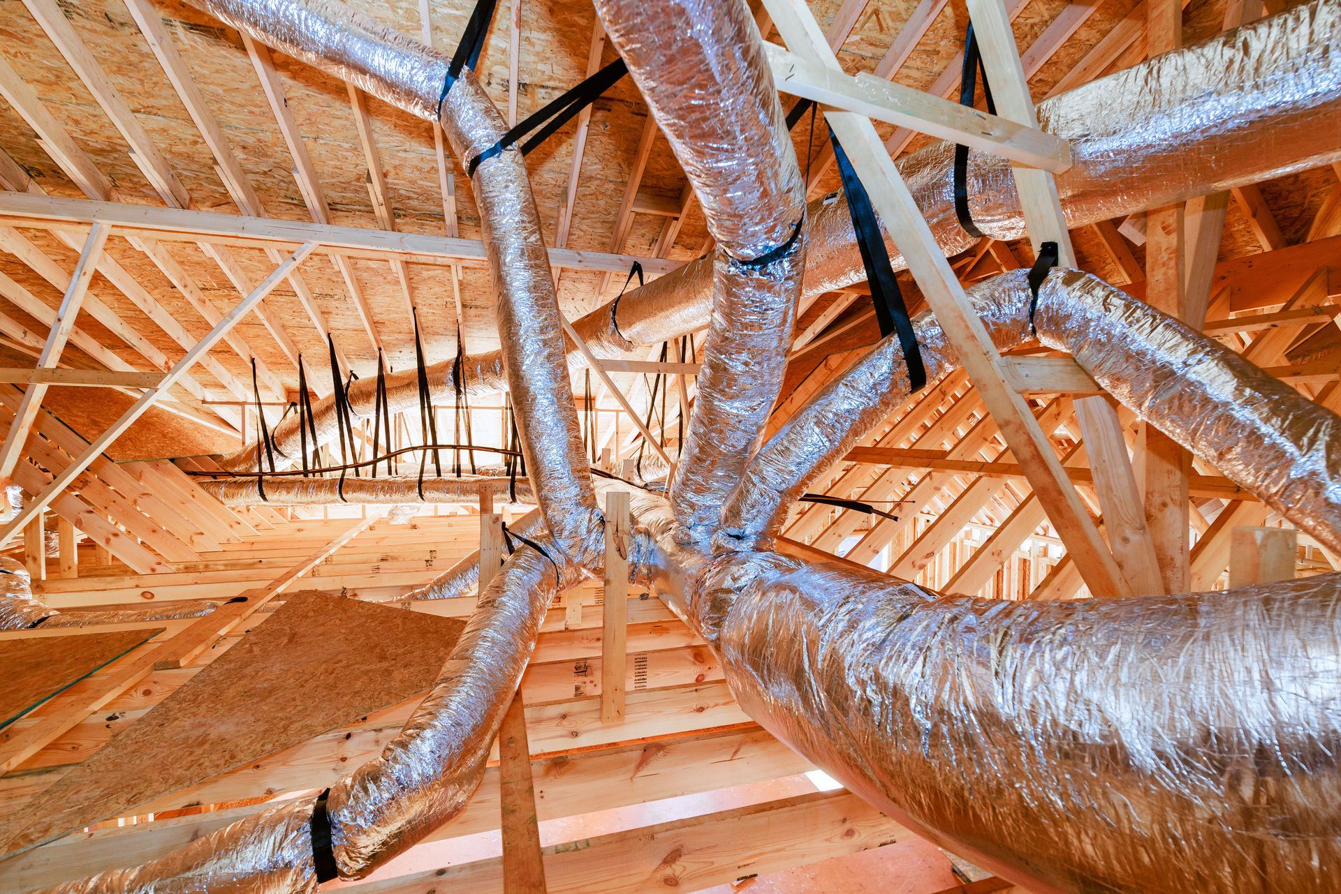 Flexible silver HVAC ducts suspended from the wooden rafters of an unfinished attic.