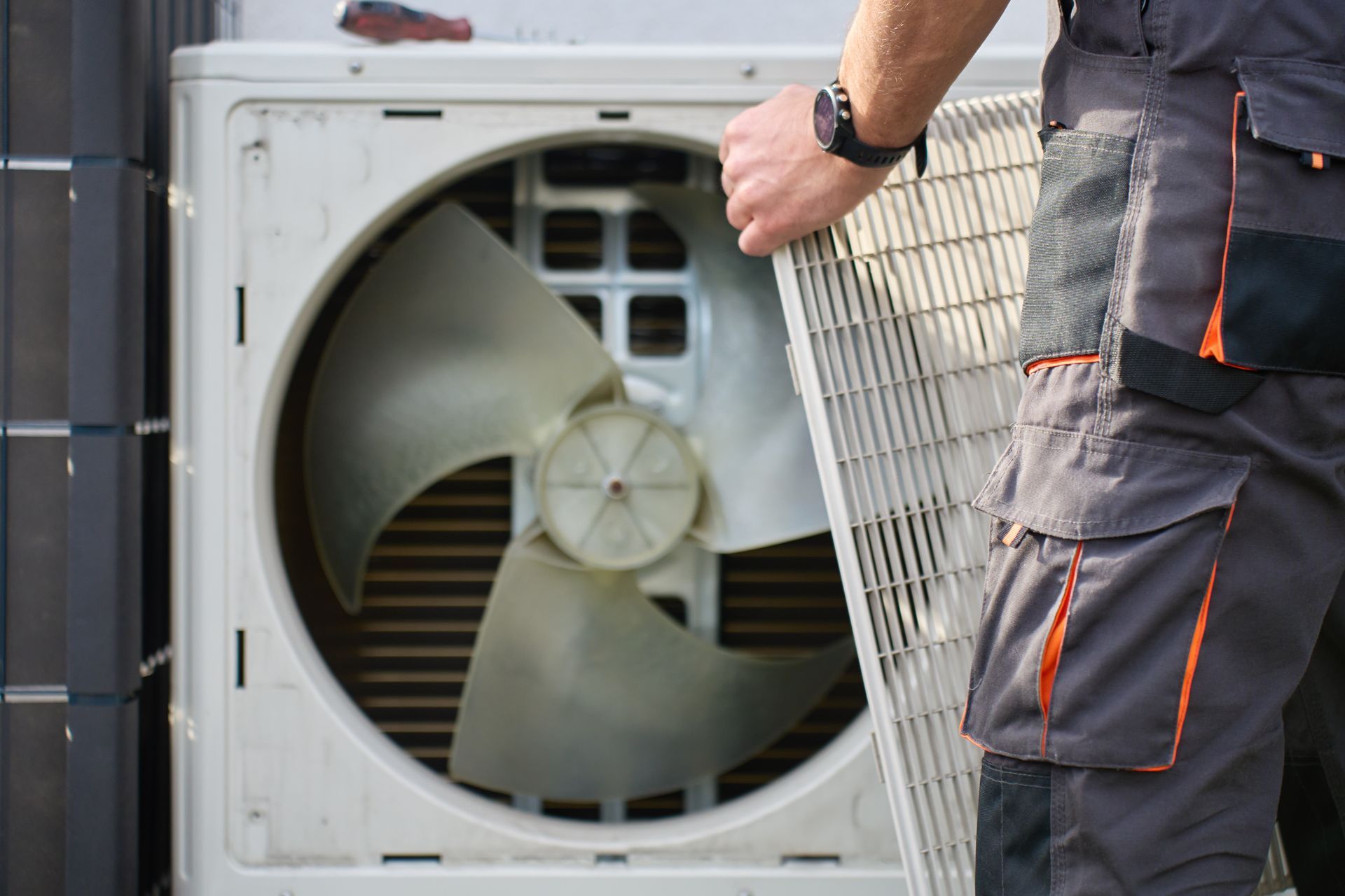 A technician in work pants holds a protective grille while performing maintenance on an outdoor air conditioning unit.