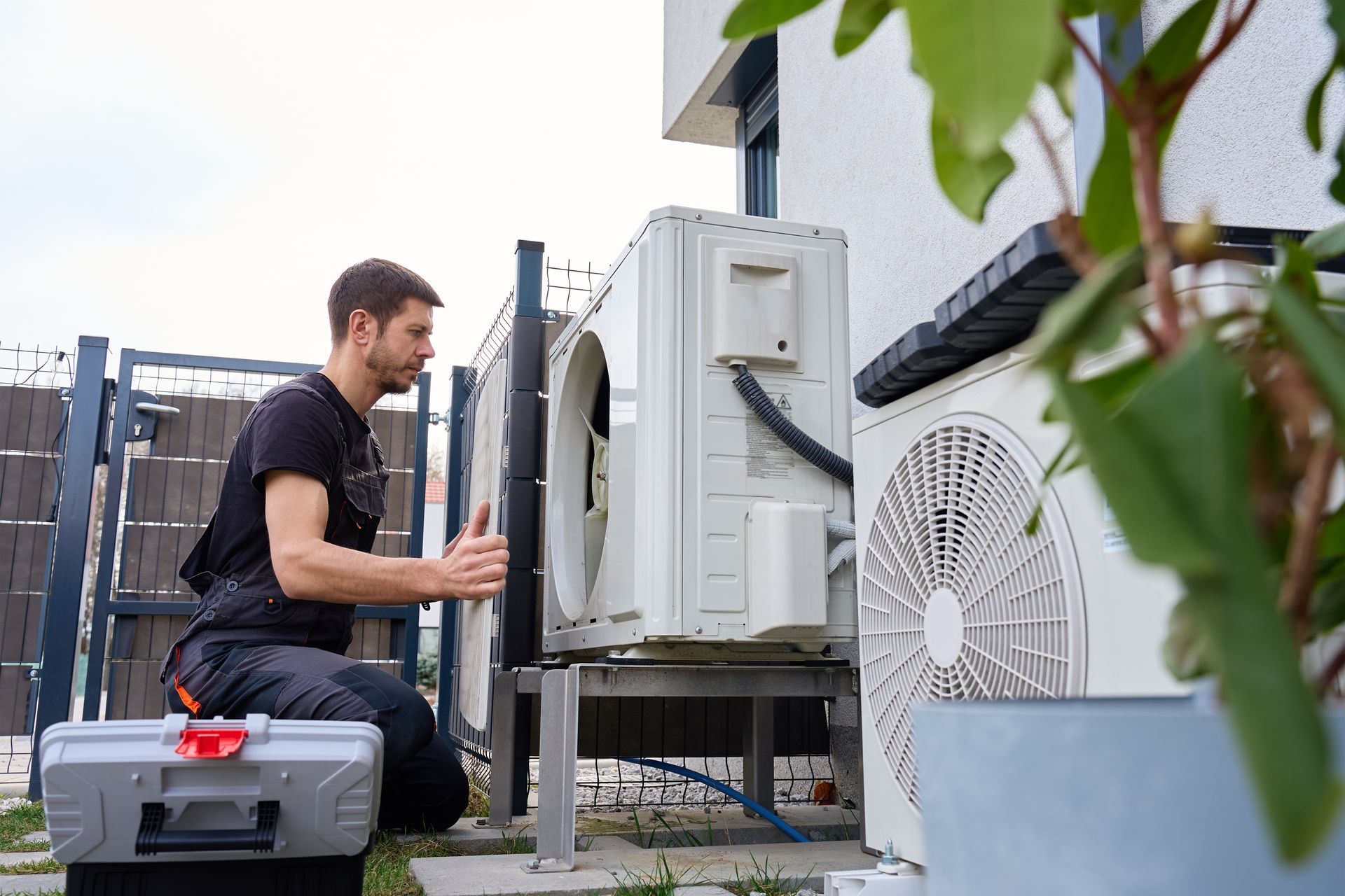 A technician in dark work clothes crouches to repair an outdoor air conditioning unit next to a building.