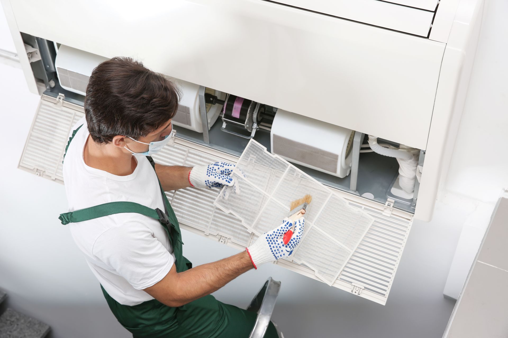 A worker in green overalls and gloves cleans an air conditioner filter with a brush.