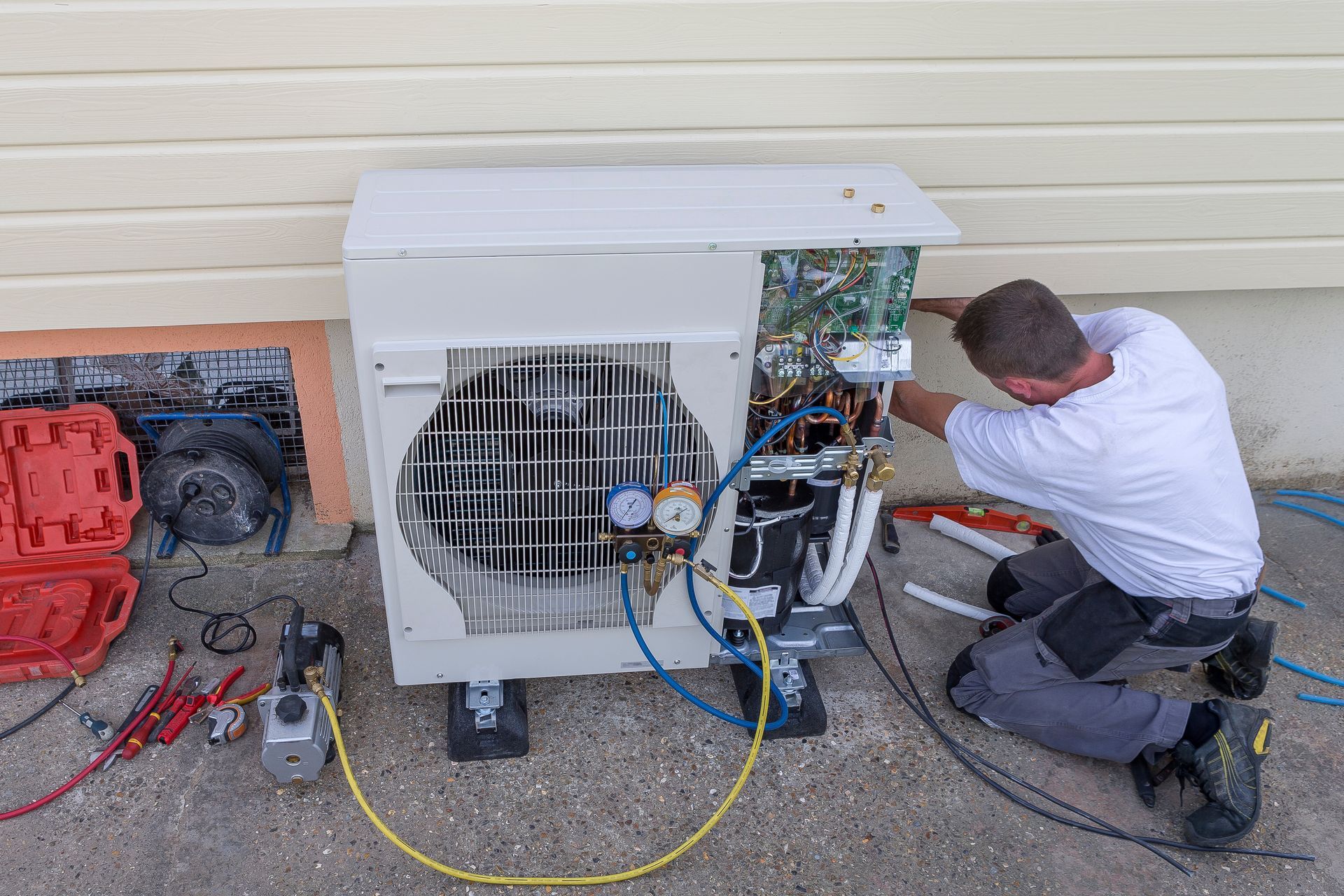 A technician repairs an outdoor heat pump unit, connecting hoses to gauges while working on a gravel surface.