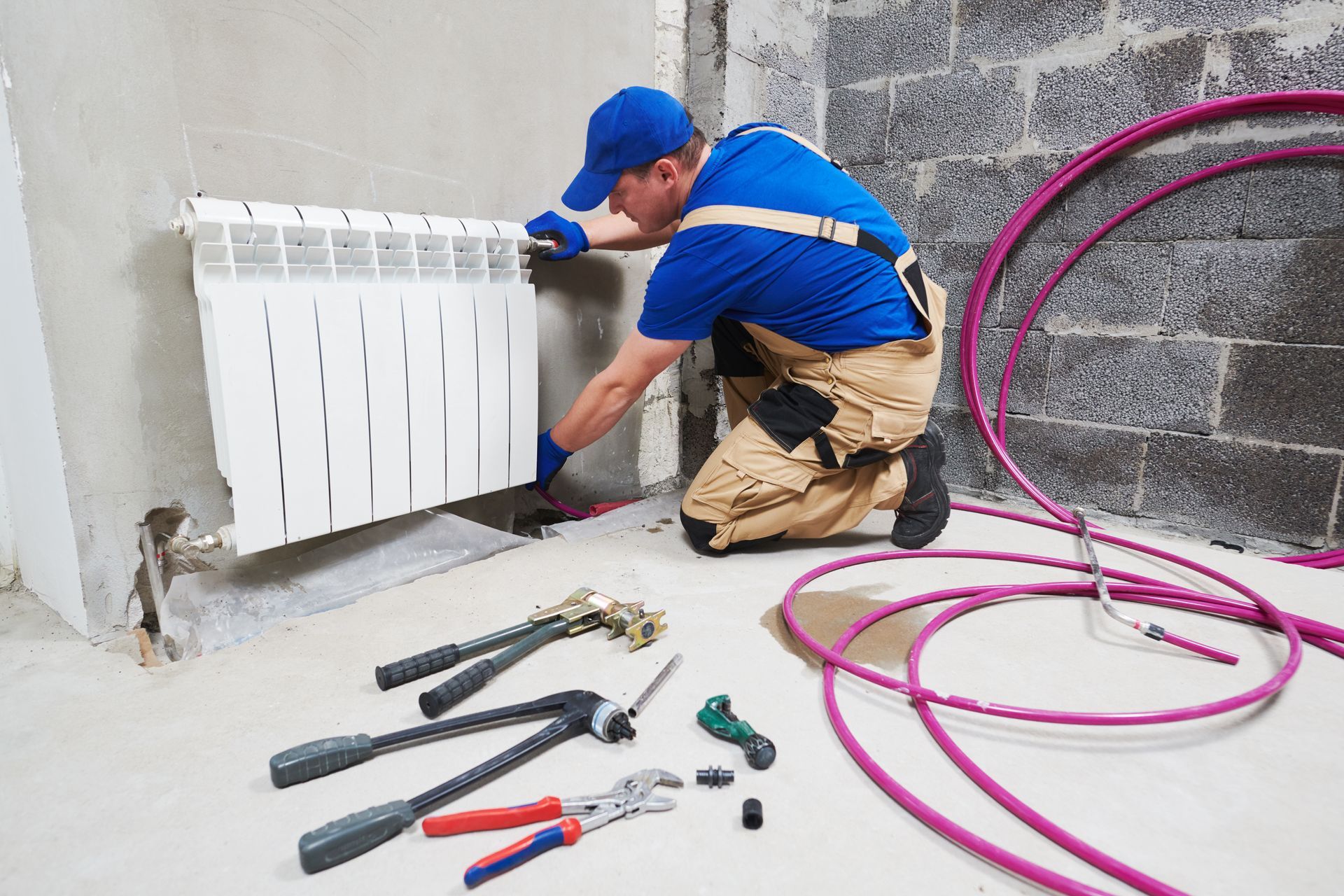 A technician wearing a blue uniform kneels on a concrete floor to install a white radiator near purple tubing and tools.