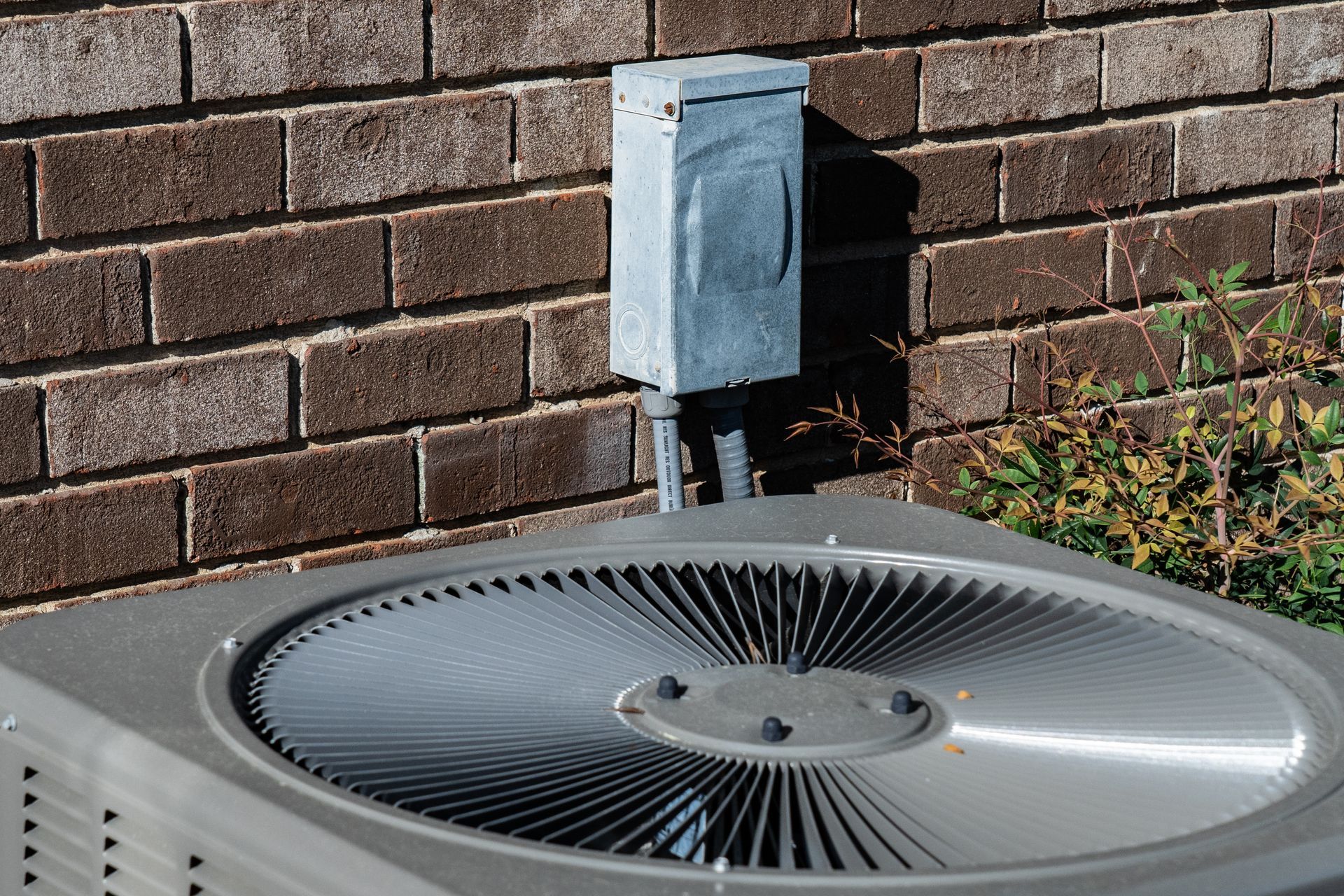 An outdoor air conditioning condenser unit next to a brick wall with an electrical disconnect box mounted above it.