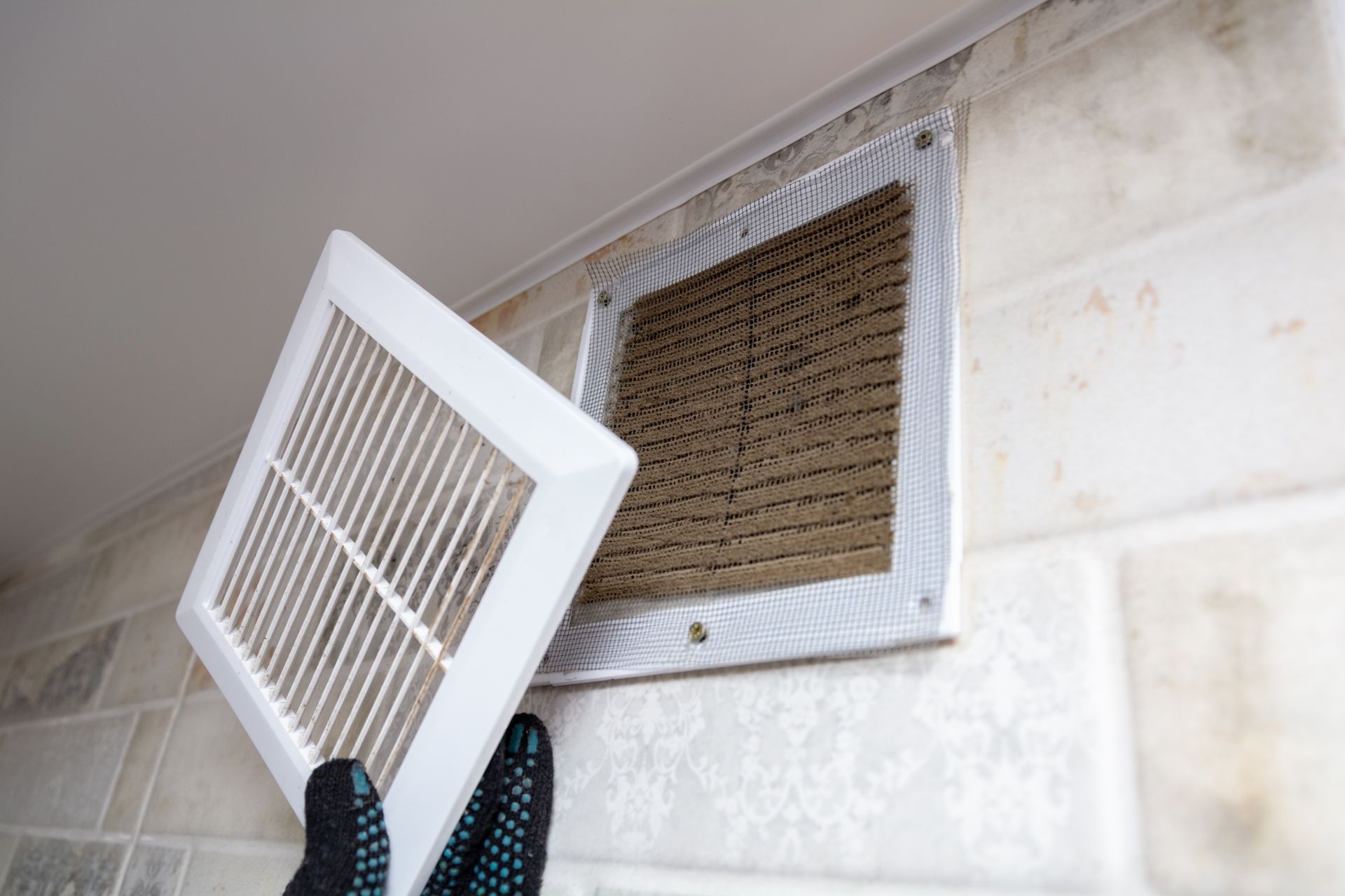 A gloved hand removes a white plastic vent cover from a wall, revealing a layer of dark dust buildup on the vent behind it.