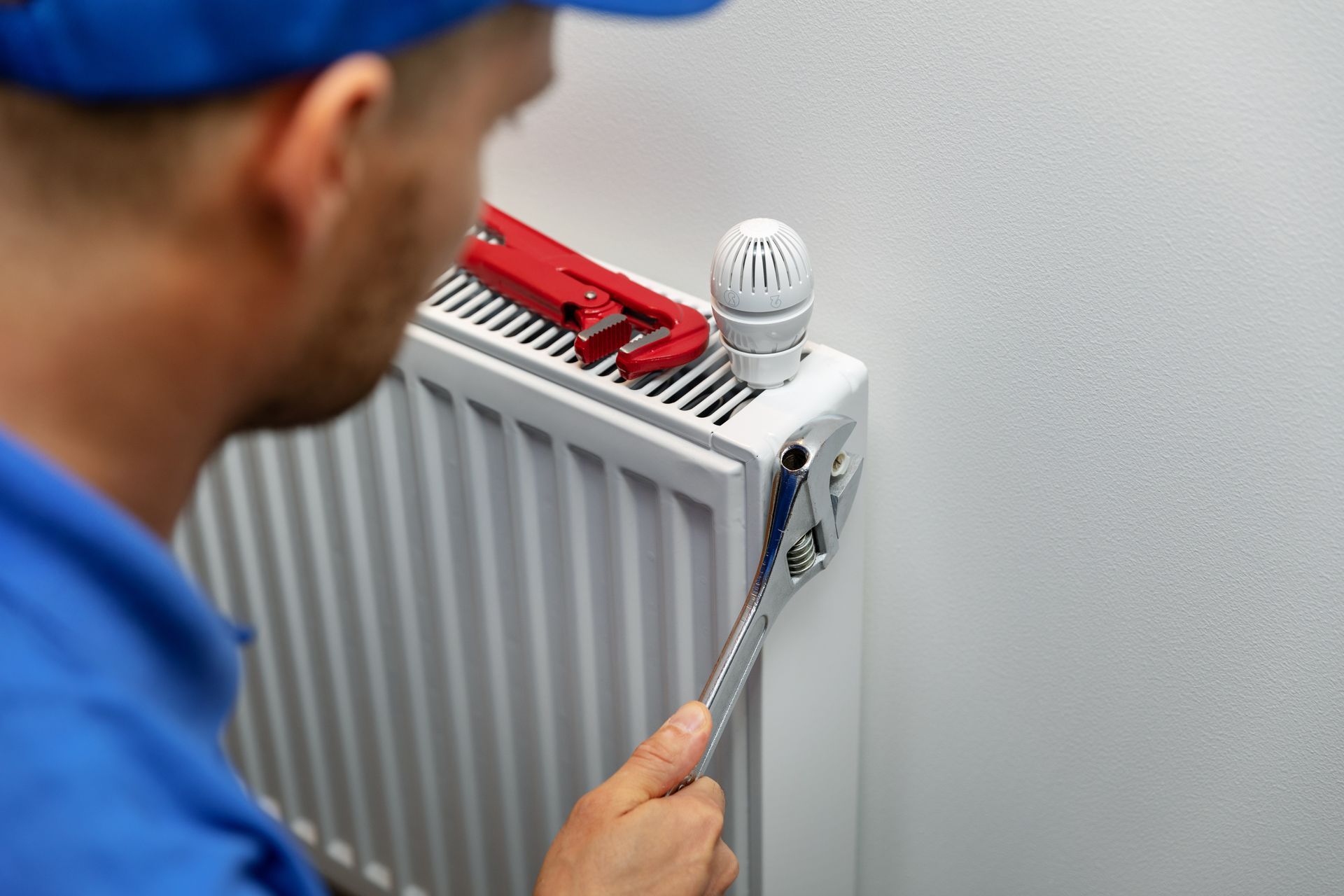 A technician in a blue uniform uses an adjustable wrench to repair a white radiator mounted on a wall.