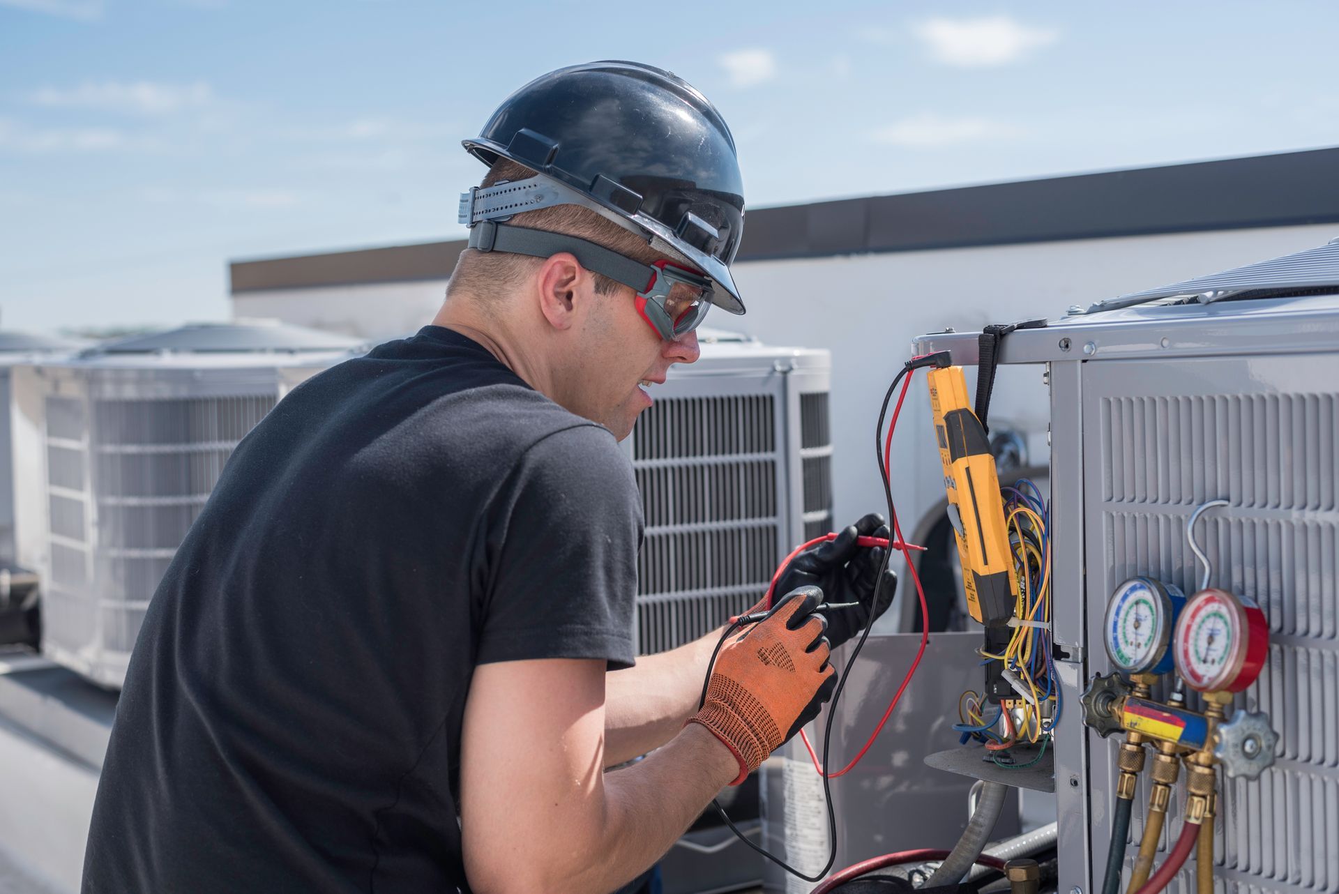 A technician wearing safety gear inspects the electrical components of an HVAC unit on a rooftop.