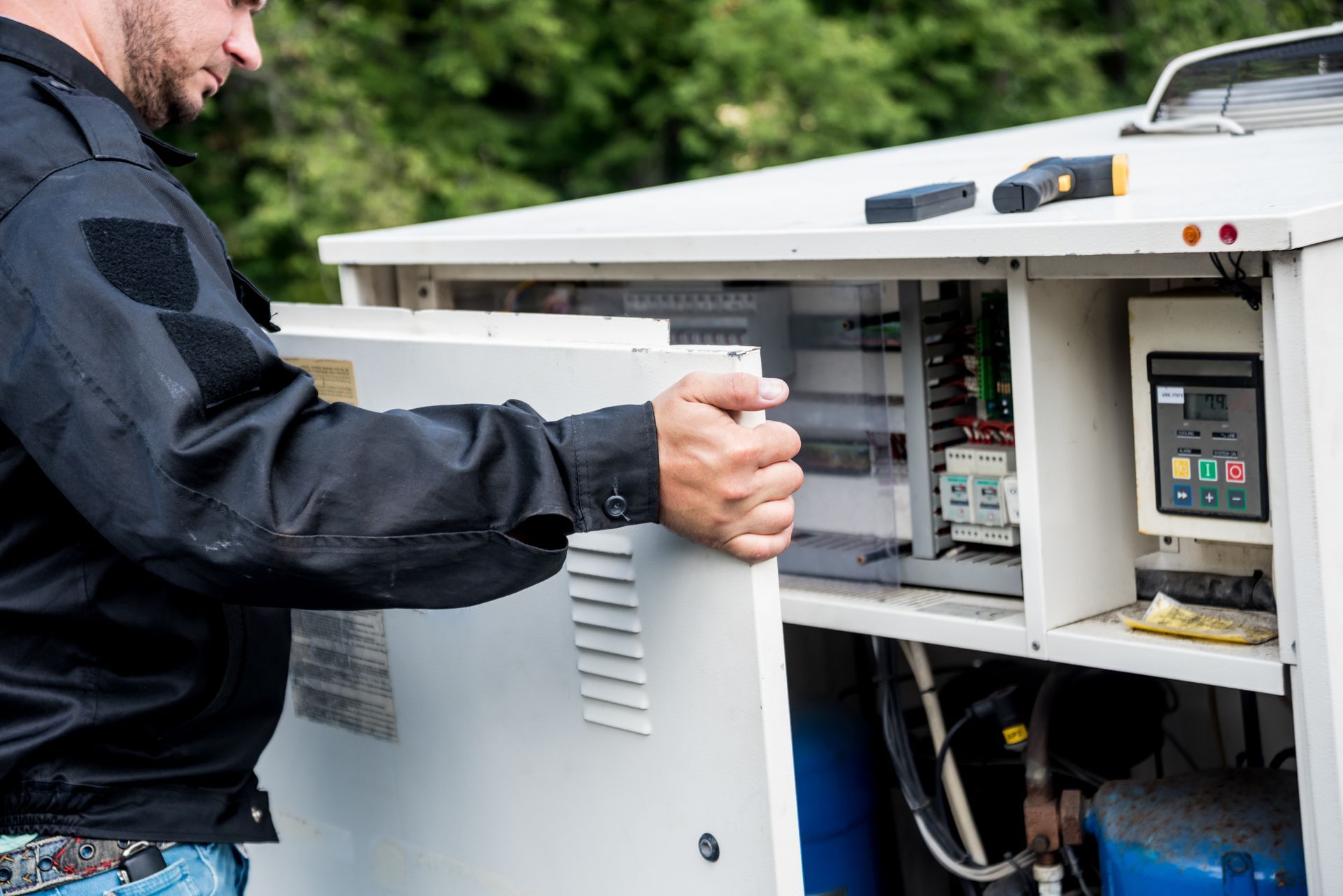 A person in black workwear opens the service panel of an outdoor industrial electrical unit.