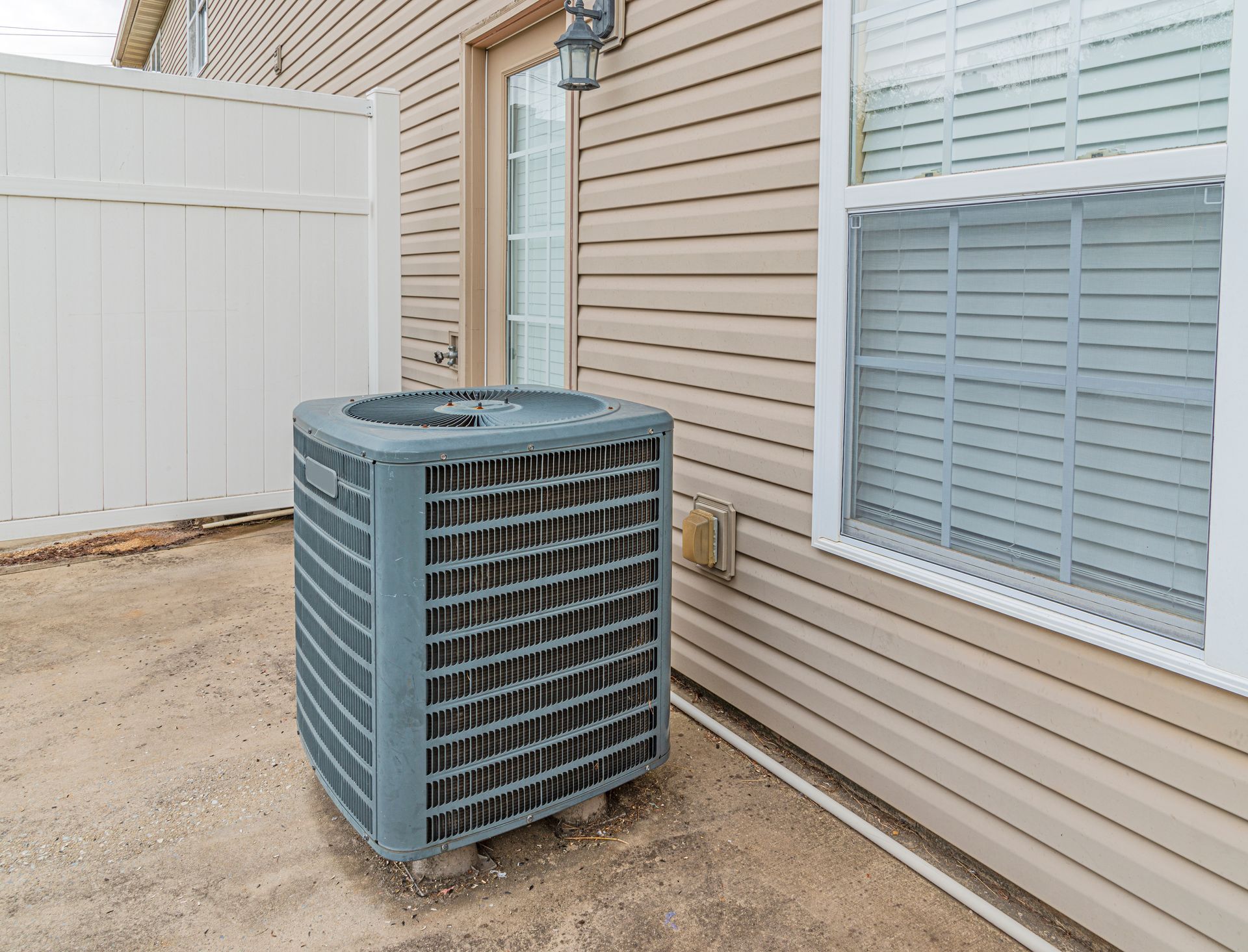 A gray HVAC unit sits outside on a concrete pad next to the tan vinyl-sided wall of a house with a white fence nearby.