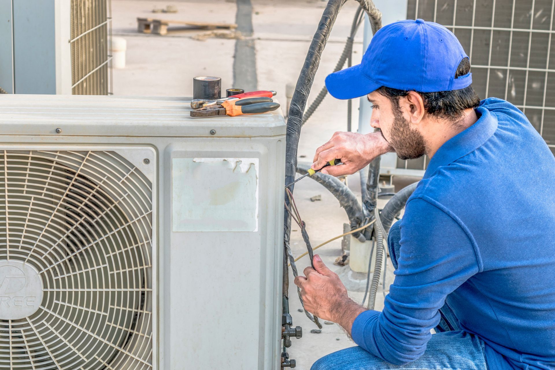 A person wearing a blue cap and shirt uses a screwdriver to repair the wiring of an outdoor air conditioning unit.
