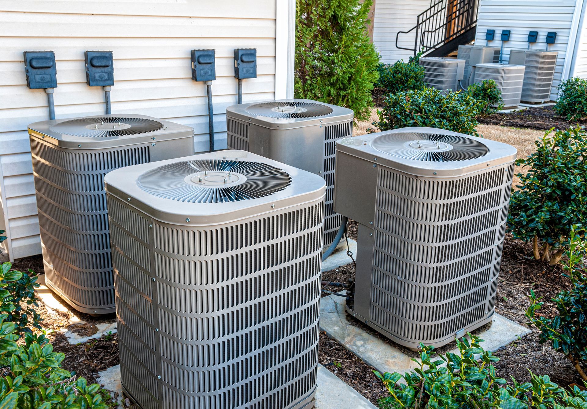 Four central air conditioning units sit on concrete pads outside a building next to a light-colored siding wall.