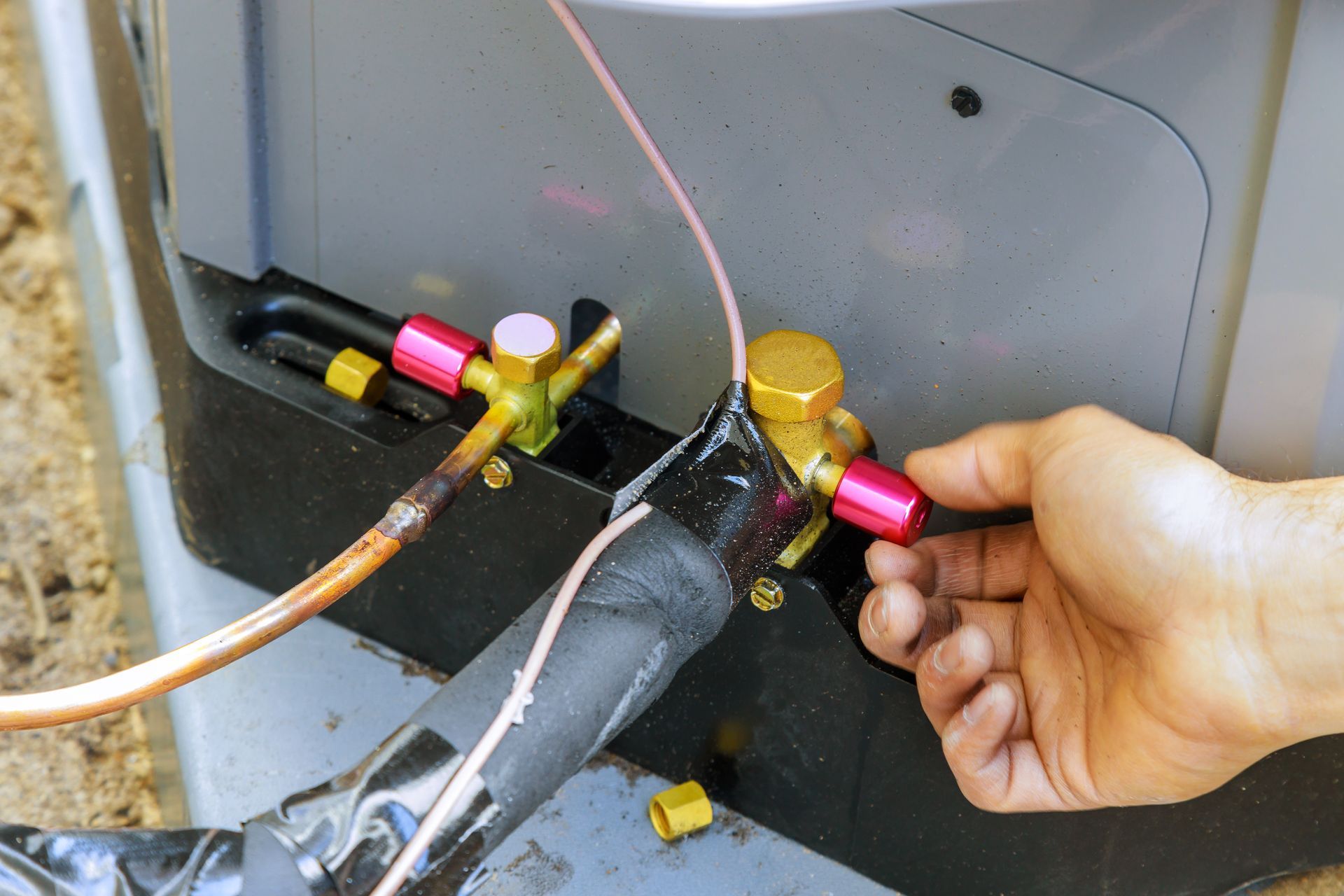 A hand unscrews a pink cap from a brass service valve on an outdoor air conditioning unit.