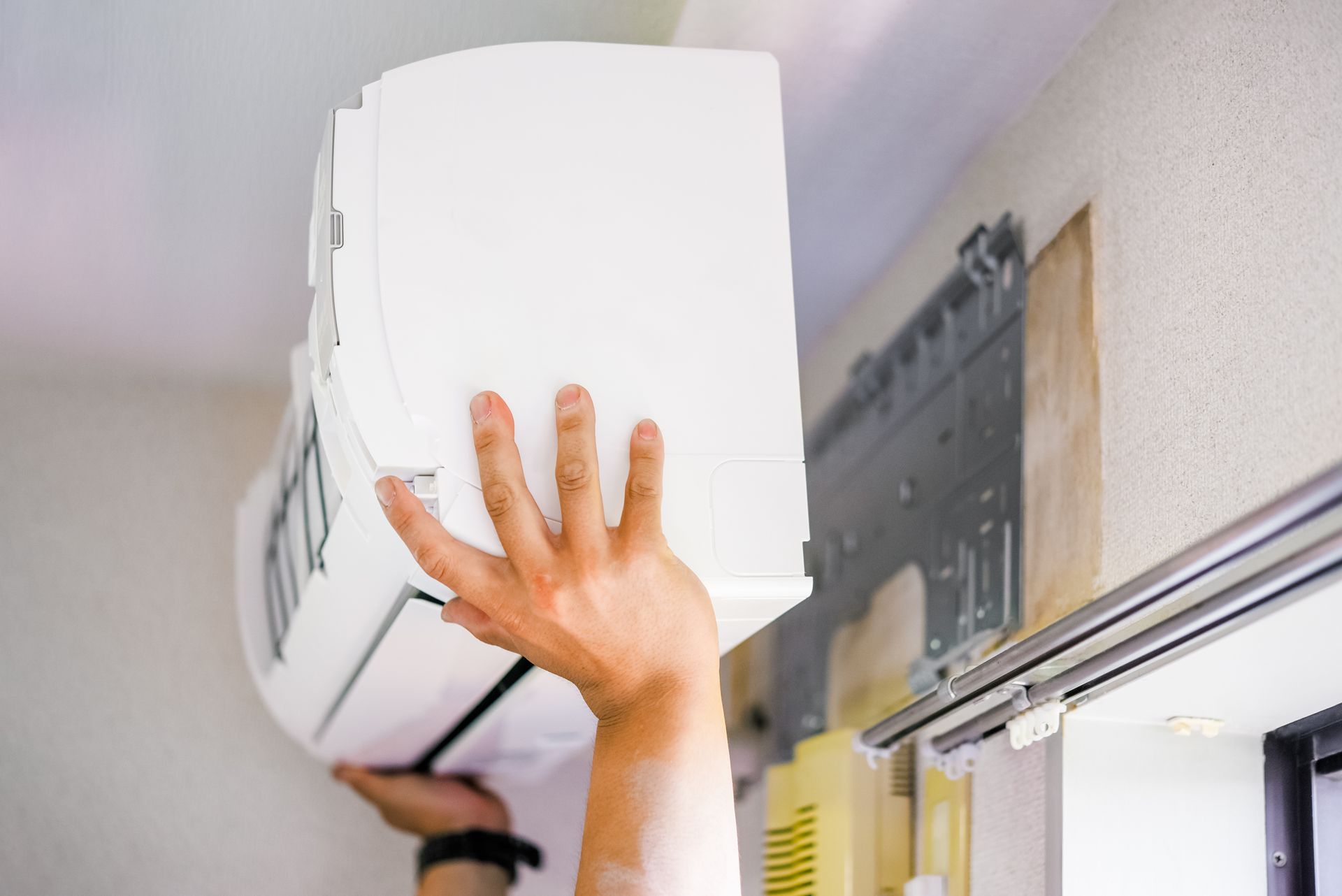 A pair of hands lifting a white split-system air conditioning unit onto its wall-mounted bracket.