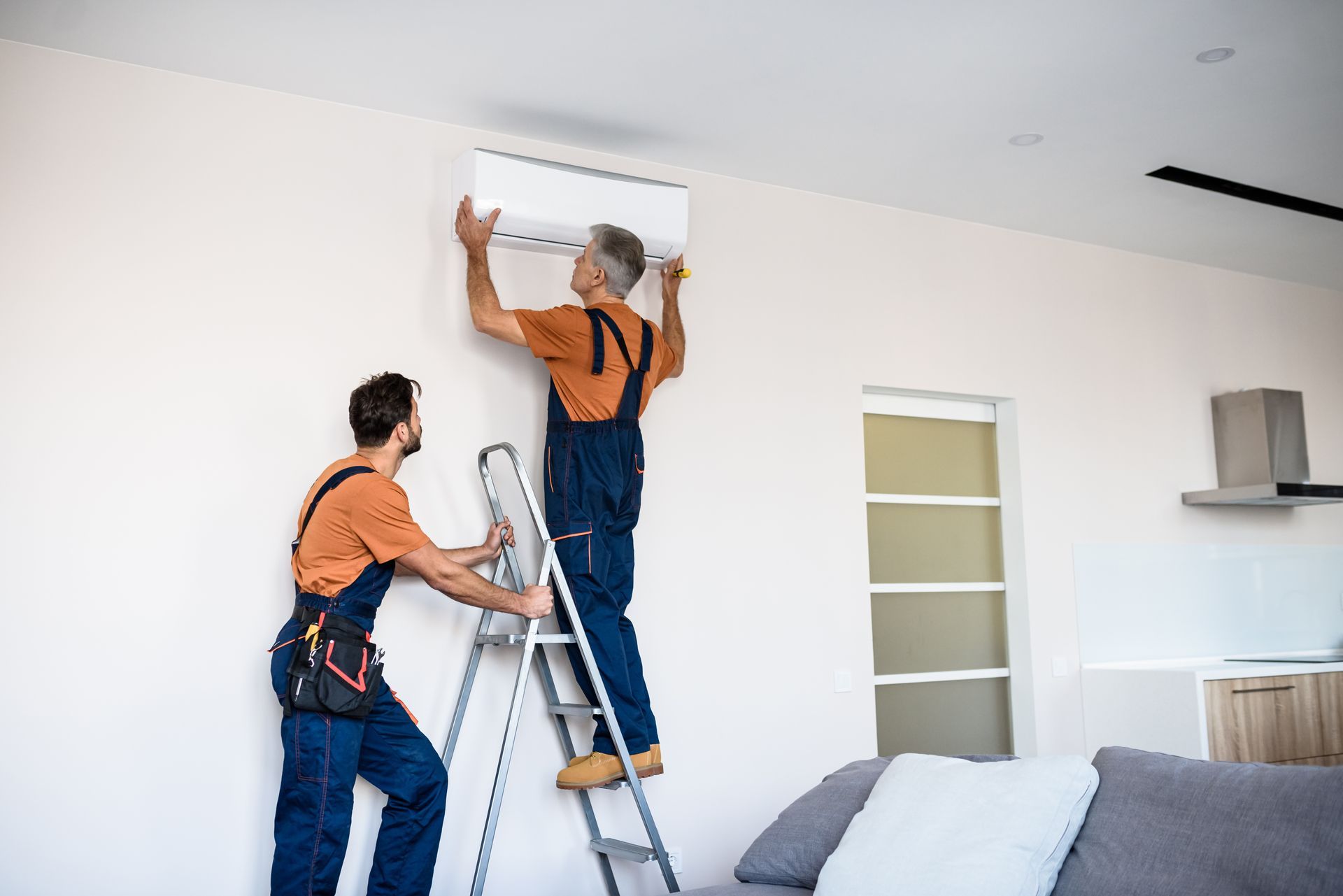 Two technicians in work uniforms installing a white air conditioning unit on a plain white wall.