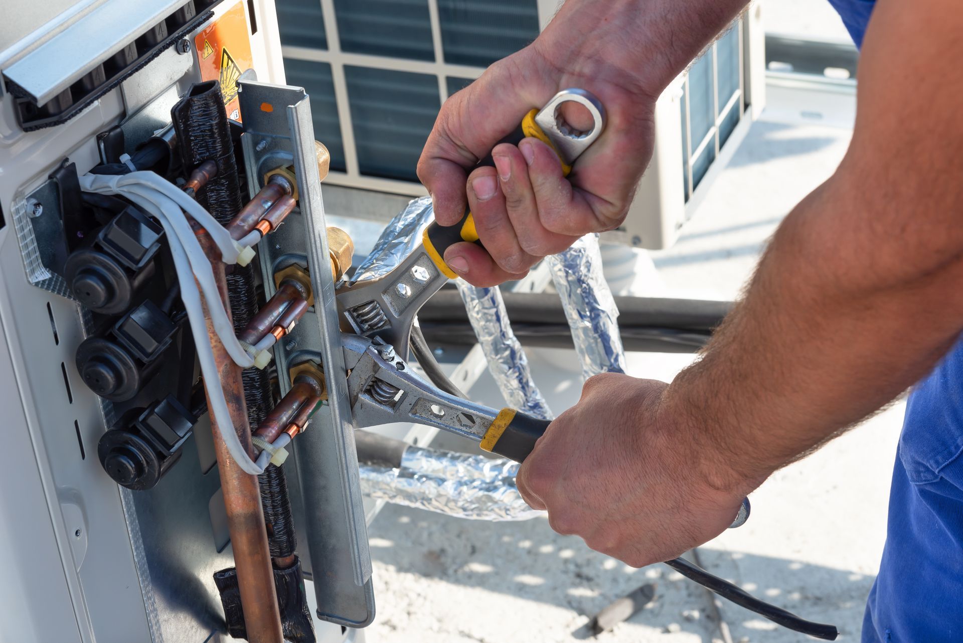 A technician uses two adjustable wrenches to tighten a copper fitting on an outdoor air conditioning unit.