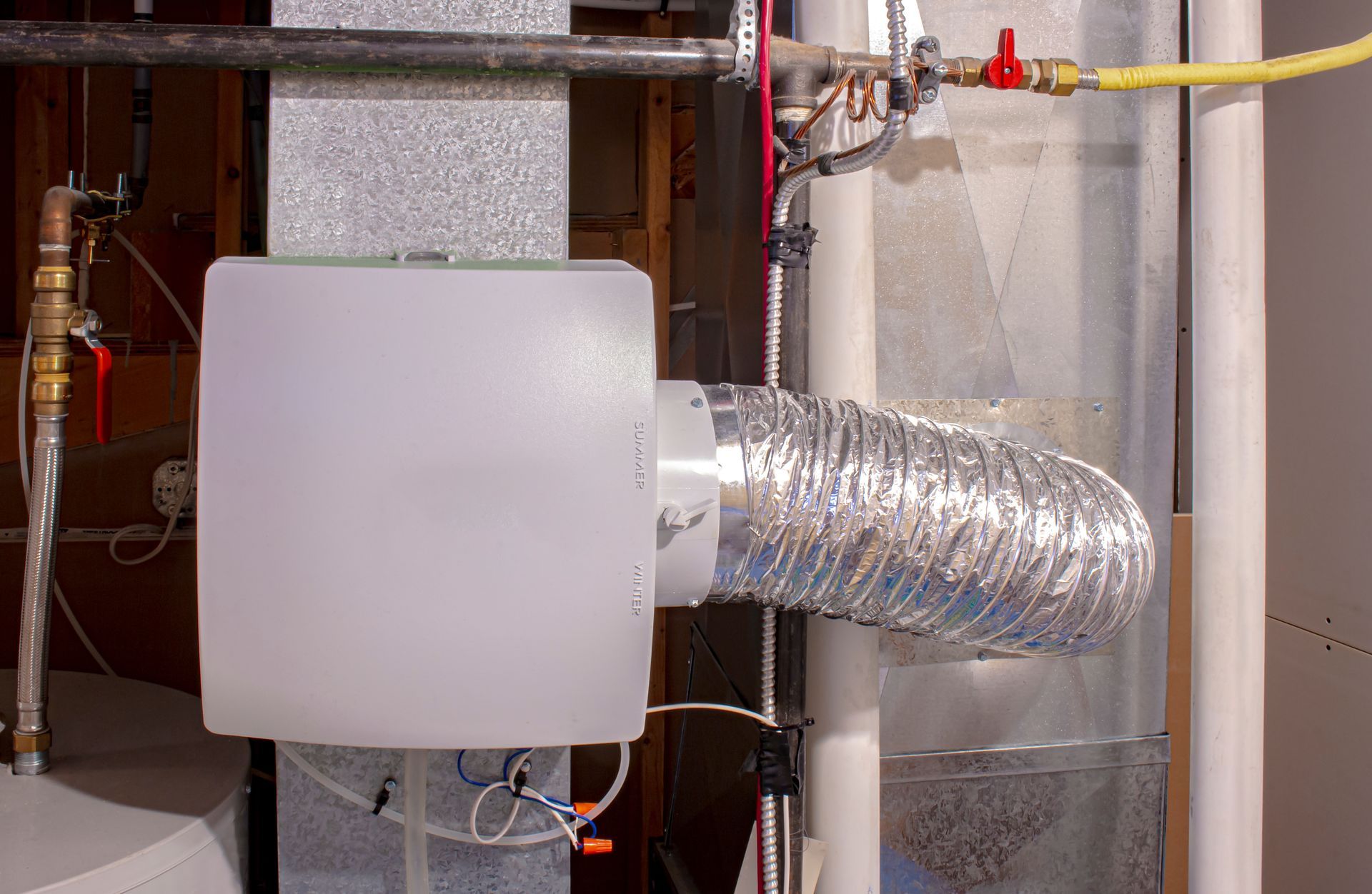 A white whole-home humidifier unit mounted to metal HVAC ductwork with a silver flexible exhaust hose.
