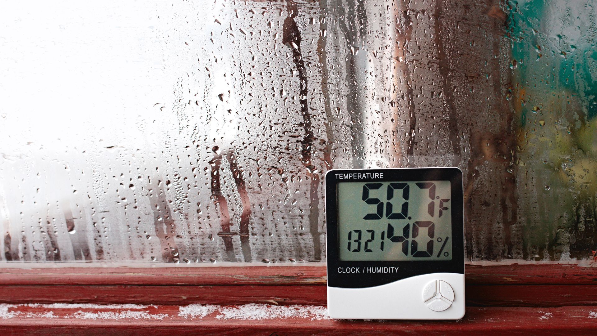 A digital thermometer and humidity gauge sitting on a windowsill covered with condensation and droplets of water.